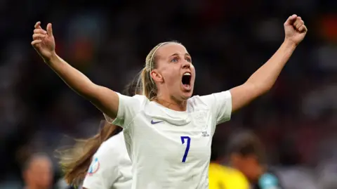 PA Media England's Beth Mead celebrates scoring their side's first goal of the game during a UEFA Women's Euro 2022 Group A match at Old Trafford, Manchester. Her long, blonde hair is tied back in a ponytail and she is wearing a white kit with the number seven on it.