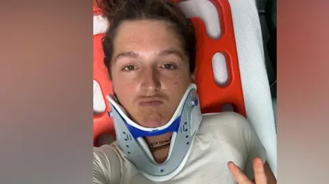Matilda Pine wears a neck brace and lies on a hospital bed. She is holding her hand up and doing the peace sign with two fingers.