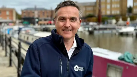 Canal and River Trust Man with grey hair smiling at the camera stood in front of a canal, wearing a Canal and River Trust navy jumper
