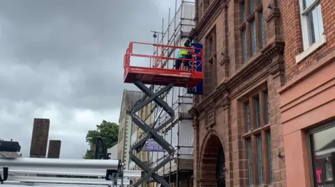 A person wearing a high-viz vest is standing on an aerial platform and is removing the sign outside Tullie.