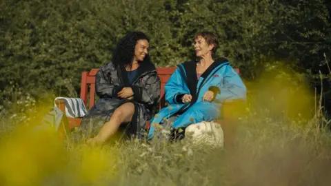 FIFTY-FOUR DAYS Cat White with long dark hair wearing a grey coat and blue top talking to Celia Imrie with short dark hair who is wearing a blue coat. They are sitting on a wooden bench on grass with bushes behind.