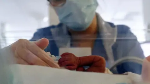 AFP via Getty Images Baby in neonatal ICU incubator, with only an arm visible. A nurse with a face mask, gloves and glasses, has her hand curled around the baby
