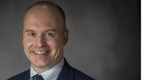 Head and shoulders shot of Andrew Crookham. He is wearing a blue suit, white and grey shirt and a blue tie. He has short hair and is smiling at the camera. He is standing in front of a grey backdrop.