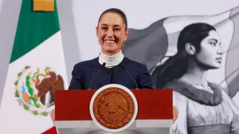 Presiden Reuters Meksiko Claudia Sheinbaum berdiri di podium di depan bendera Meksiko dan mural hitam-putih yang menunjukkan seorang wanita mengibarkan bendera. Presiden Sheinbaum tersenyum luas sementara dia mengadakan konferensi pers di Mexico City.