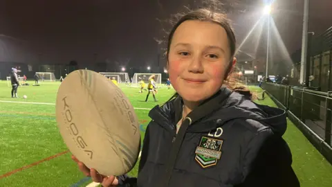 Elizabeth Baines / BBC A girl with brown hair tied up wears a black puffer coat. She stands on a pitch and holds a rugby ball