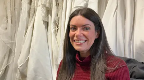 A woman with long brown hair wearing a red jumper smiles at the camera. She is sitting in front of a row of white wedding dresses