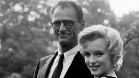 PA Media Arthur Miller stands beside Marilyn Monroe in a black-and-white photograph, both facing the camera with trees visible in the background.