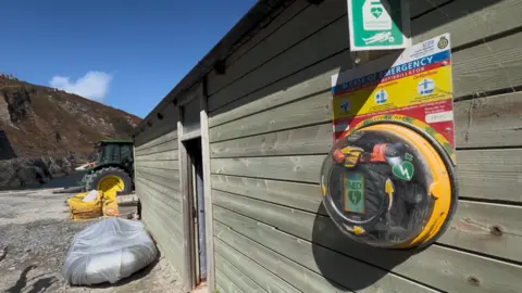 Tara McEvoy-Wilding A defib is attached to the wooden wall of a boathouse. It has a green sign which says defibrillator and there is a yellow, red and blue emergency notice telling people what to do.