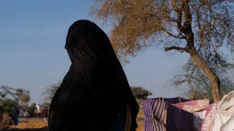 An unidentified woman wearing an abaya and hijab walks through a camp in Chad. Her face is not visible.