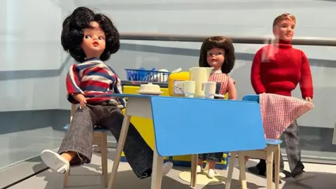 A Sindy doll sits at a 1960s-style table with a smaller female doll and a male doll