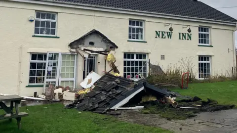 BBC Front of New Inn pub with lettering attached to wall of long building painted cream colour. The front porch has been destroyed and debris is strewn across the path leading to the entrance.
