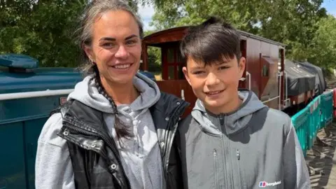 A woman with dark hair in a pigtail, with a hint of grey at the front, is wearing a grey hoodie and a black open-zipped gillet. She is standing next to her son, who is in his mid-teens and has a grey hooded top on. He has short black hair. They are both smiling into the camera and in the background is a locomotive train.
