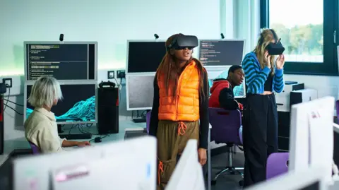 Students in a classroom with computers. Two female students are wearing VR headsets. One student wears an orange gilet and the other a blue stripy cardigan. Behind them are students looking at them and the computers, which show code on the screen.
