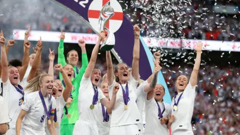 Reuters A group of female England football players lift the trophy on the pitch as confetti falls. 