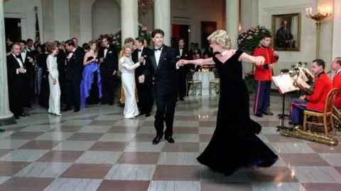 Pete Souza/The White House via Getty Images Princess Diana in long black dress dances with John Travolta in the White House. They are watched by a small crowd and there's a band to their right. 