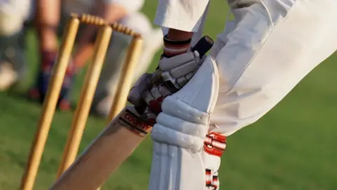 a close-up of a cricketer wearing cricket whites holding a cricket bat with gloved hands, standing in front of a set of wooden wickets. A blurred player can just be seen crouching down behind the wickets.