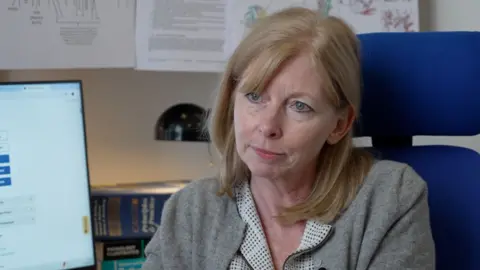 A white woman, with fair, shoulder-length hair sits in a blue office chair. She is wearing a checked shirt and grey cardigan. On the desk behind here there is a computer monitor, books and a lamp.