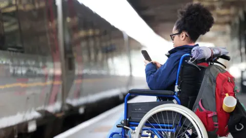 Female wheelchair user waiting on a train platform, using her phone.