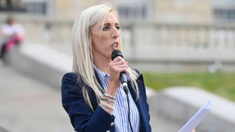 PA Media Carla Lockhart addreses an audience with a microphone in one hand, and a sheet of paper in the other. She is wearing a navy blazer, with a blue and white striped shirt