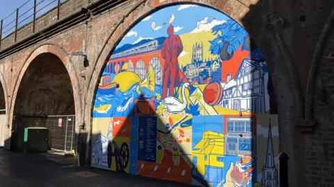 A bright-coloured mural decorated onto an archway under a railway bridge. The mural is made up of mainly, yellow, blue and orange pictures. The arch to the left of the mural is yet to be redeveloped and remains as open red-brick. 