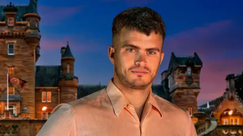 Traitors contestant Matthew Hyndman, with short brown hair and stubble, wearing a light pink shirt. He is standing in front of the Traitors castle and it is nighttime. 