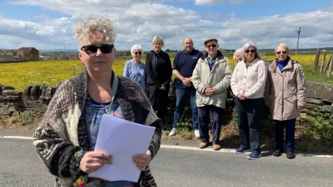 A woman with short silver hair and wearing sunglasses standing in front of a group of people next to a dry stone wall