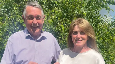 The Souttars in blue and white shirts standing side by side smiling in front of a tree