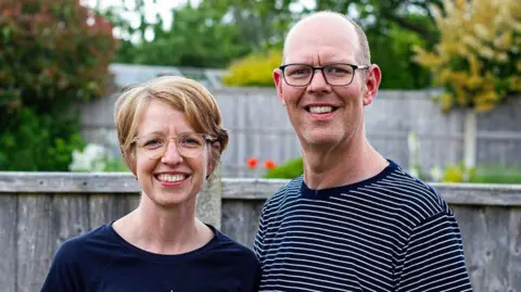 A smiling Chris Marriott with his wife Bryony stands in front of a wooden garden fence in a striped T-shirt and glasses