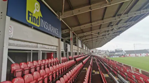 Rows of raked red seats form the stand at Celtic Park. Against the rear wall of it is a sign for Find Insurance, which is dark blue and alien green. Some of the grassy pitch can be seen. 