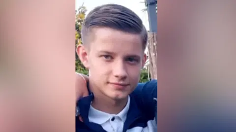A close-up photograph of Ronnie Tillot. The teenager is wearing a buttoned-u[ white polo shirt with a collared jumper over the top. His light brown hair is neatly combed to one side. Someone who is out of shot has their arm around his shoulders.