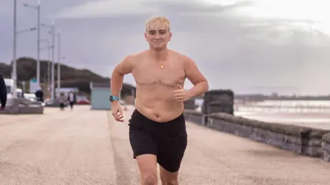 Hana Laurie Sam Smith running topless down a promenade beside a beach on a cloudy day. She is wearing black running shorts, a gold necklace and a blue smart watch. She has short blonde hair and is smiling at the camera.