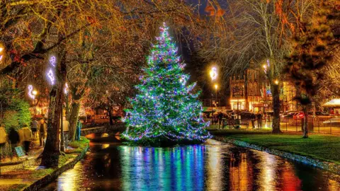 David Hanks A Christmas tree in the river in Bourton-on-the-Water lit up at night. The tree's colourful blue, green and pink lights are reflected in the river, while yellow, orange and red lights from near by shops are also picked up in the water. People are walking along the river bank and there is a small low bridge behind the Christmas tree. 