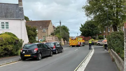 Police cordoned off the area at Fordington High Street where the gas leak was found. A policeman and a resident can be seen. Several cars are parked nearby. It is cloudy.