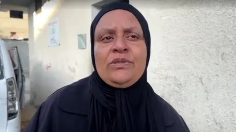 Reuters Umm Muhammad Abu Aisha, wearing black clothes and headscarf, with white walls and a car behind it