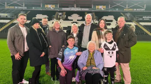 A group of people standing on a grass pitch smiling into the camera. There are several men and women in coats and two boys standing at the front with an older women sitting in a wheelchair.