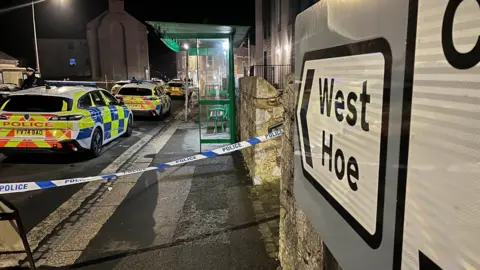 BBC A West Hoe sign on the right with police tape across a pathway. There are several police vehicles behind the police tape, along with a bus stop on the pathway. The photo was taken at night so the pathway and road is lit up by street lights.