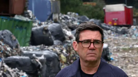 Burnham looks on at an illegal rubbish dump near the Bradford City football ground. Red, green, blue and brown skips are piled on top of black bin bags and mountains of loose rubbish in the background. In the foreground, Burnham looks serious and is wearing a navy raincoat over a black t-shirt, with black spectacles and short black hair.