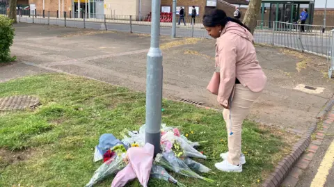 Ghuilermina, a woman looking down at a collection of flowers that have been placed around a lamp post. She is wearing a pink top, pink trousers, white trousers and has long dark hair. There are metal railings in the distance. 