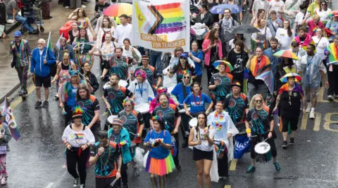 Steve Brock Photography A group of people at a pride march. Many are wearing bright colours, some wearing blue NASUWT t-shirts. The weather is raining and some are carrying umbrellas. There is a banner with one of the pride flags and the logo "coming out for teachers".