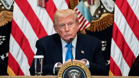 US President Donald Trump, in a dark suit, sits behind a desk at the White House with the presidential seal in front of him and a flags, including the Stars and Stripes, behind him
