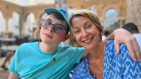 Family photo Teenage boy, Lucas, wearing a blue t-shirt, blue hat and black glasses with his arm around his mum, Louise wearing a blue and white blouse.