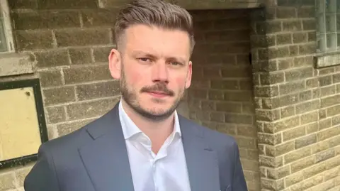 LDRS A man with short dark hair wearing a white shirt and grey suit jacket.