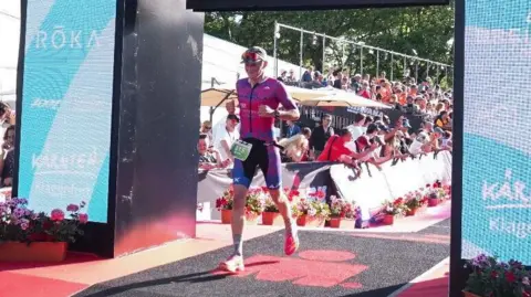 Braden Morris passing over the finish line. He is wearing blue and pink running gear and a cap with sunglasses.
