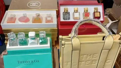 LDRS Fake Chanel and Tiffany perfume in boxes on a table along with a fake Marc Jacobs tote bag.