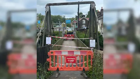 Alex Wilson A suspension bridge with a 'footpath closed' sign and barrier in front of it. The bridge has red and white tape on it as well as some white signs. On the opposite side of the bridge is some scaffolding.