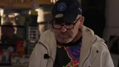 Lawrence Cook, wearing a black cap, beige coat and black-rimmed glasses, looks at the floor in his darkened flat