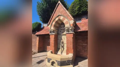 A fountain surrounded by bricks with trees and blue sky in the background