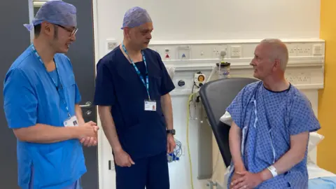 A surgeon wearing blue scrubs, a surgeon wearing navy scrubs and a patient wearing a blue gown. The surgeons are wearing blue head covers. The patient is sitting up in a bed.