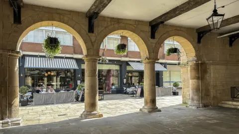 View across the Square in Shrewsbury town centre from the Old Market Hall. There is a paved area and the building has arches and pillars. Across the square, people are sitting at tables and there are shop awnings.