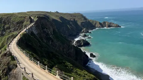 A coastal cliffside footpath curves along rugged headlands above turquoise water, with waves breaking against rocky outcrops below under a clear blue sky.